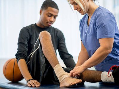 A male teenage basketball player is at the doctors office to have some work done on his injury. A nurse is wrapping his bandage around the ankle.
